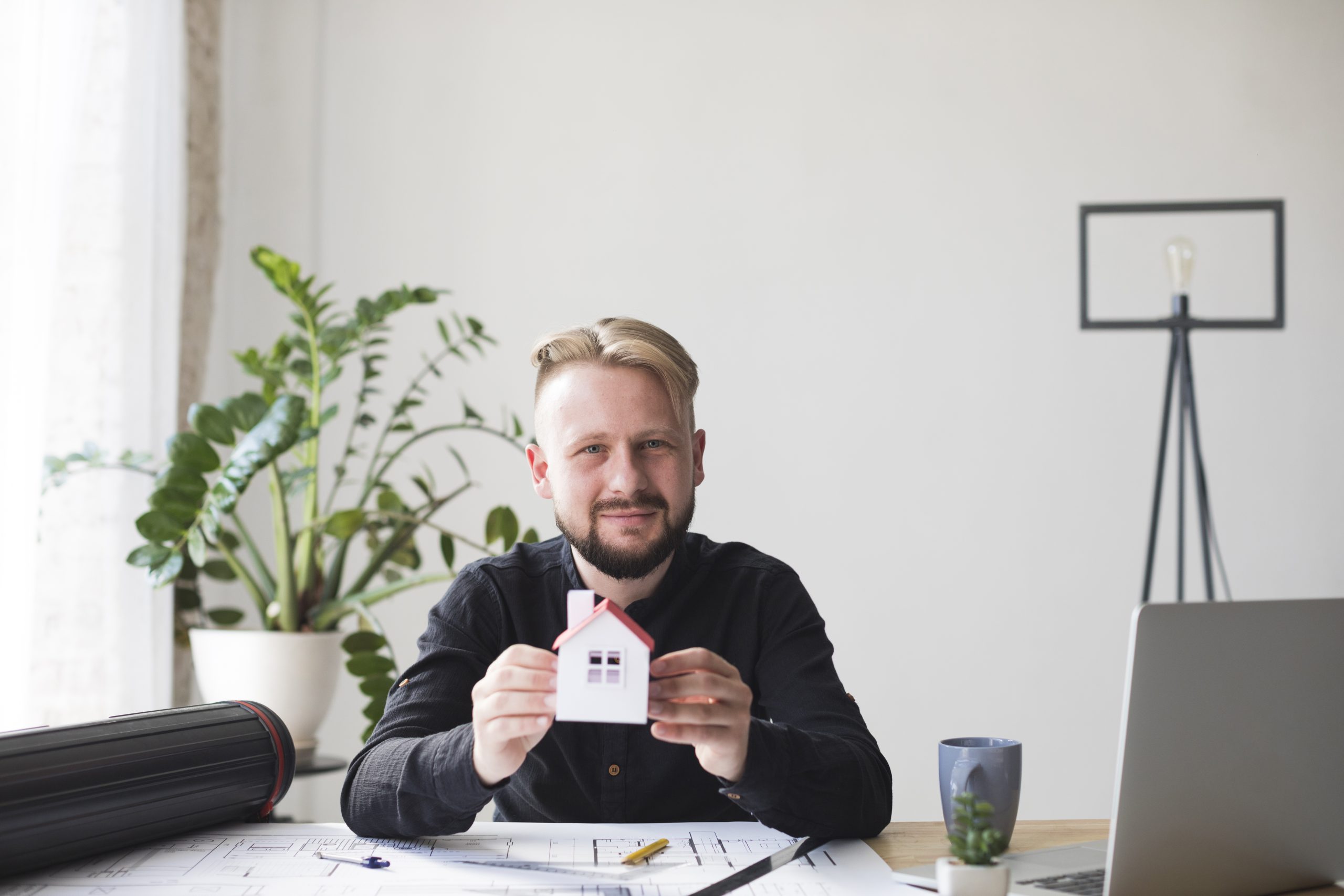 portrait-smiling-young-man-holding-house-model-sitting-office-looking-camera